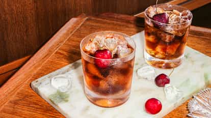Dos cócteles Black Russian con hielo y cerezas en vasos bajos sobre una bandeja de mármol, rodeados de cubos de hielo y cerezas frescas.