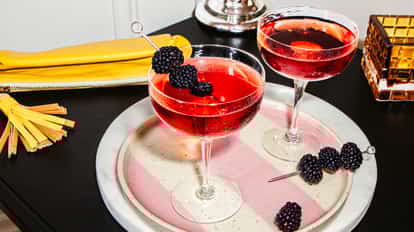 Two Blackberry Martini cocktails in coupe glasses with fresh blackberry garnishes on a pink striped tray beside a yellow clutch.