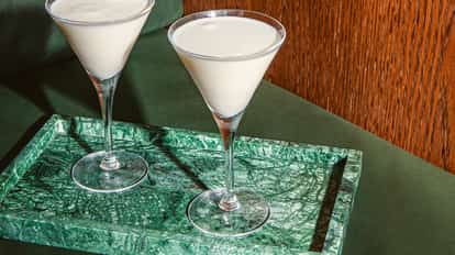 Two Blanc Martini cocktails in tall martini glasses on a green marble tray, set against a green sofa and warm wood paneling.