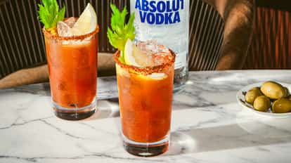 Two Bloody Mary cocktails with lemon, celery, and spiced rims on a marble table beside a plate of green olives and a vodka bottle.