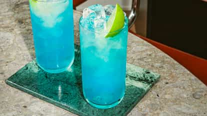 Zwei hohe Gläser des Blue Andy Cocktails, leuchtend blau mit Eis und Limette auf einer Steinplatte.