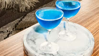 Blue Dolphin Martini cocktails served in chilled coupe glasses with vibrant blue color, set on a white marble tray on a wooden table beside a textured rug.