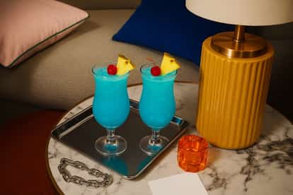 Two Blue Hawaii cocktails  on a table next to a lamp.