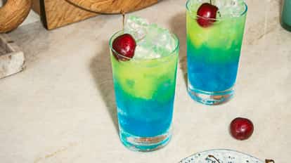 Blue Hawaiian cocktails in tall, chilled glasses with layered blue and green tones, topped with ice and cherries, set on a light marble countertop.