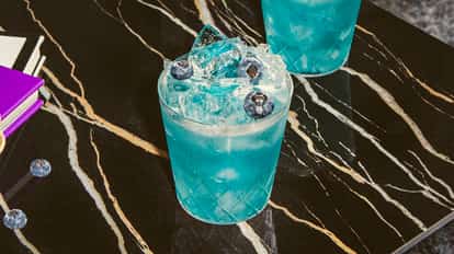 Cóctel Blue on Blue en un vaso texturizado con hielo triturado y arándanos frescos, servido sobre una mesa de mármol negro.