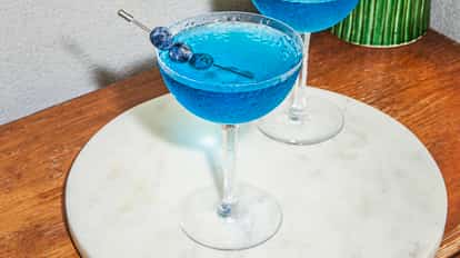 Chilled Blueberry Martinis in coupe glasses with bright blue color, garnished with blueberries on a pick, set on a round marble tray on a wooden surface.