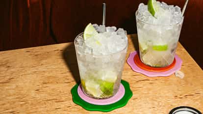 Caipiroska-Cocktail in zwei eisgefüllten Gläsern mit Limettenstücken und Metallstrohhalmen auf bunten Untersetzern auf einem hellen Holztisch.
