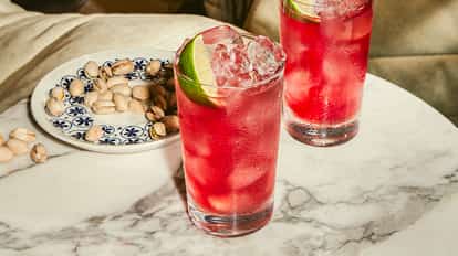 Zwei hohe Gläser Cape-Cod-Cocktail mit Wodka, Cranberrysaft, Eis und Limettenspalten auf Marmortisch, Pistazien auf kleinem Teller.