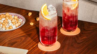 Two tall glasses of Cosmow Mule cocktail with ice, cranberry juice, ginger beer and orange slices on a wooden table, served with caramel popcorn.