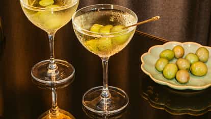 Two dirty vodka martinis with green olives in coupe glasses on a dark reflective table beside a plate of fresh green olives.