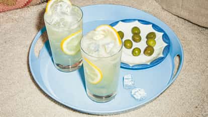 Cócteles Elderflower Collins con rodajas de limón y hielo en una bandeja azul junto a un plato de aceitunas verdes sobre una alfombra.