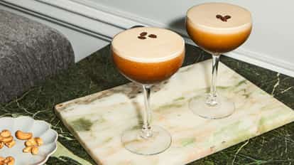 Two espresso martinis in elegant cocktail glasses on a marble table with roasted cashew nuts on a small plate.