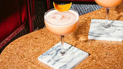 Fancy Nancy cocktail in a coupe glass with creamy foam and orange peel garnish on a cork table, elegant bar setting with marble coasters.