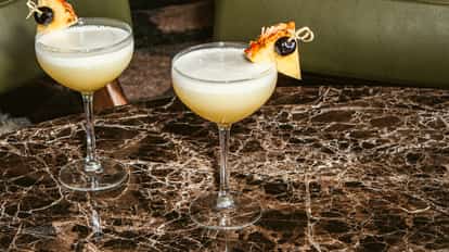 Drinks Flirtini em taças coupe com abacaxi e cereja como guarnição, sobre uma mesa de mármore escuro.