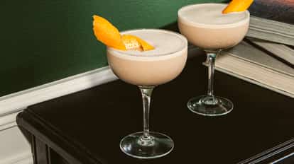 Cócteles Galway Grey en copas coupe frías de tono beige cremoso, decorados con piel de naranja, colocados sobre una superficie oscura junto a libros apilados.
