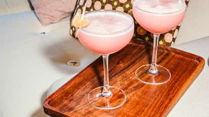 Ginger Cosmos in coupe glasses with candied ginger garnish on a wooden tray over a light sofa.