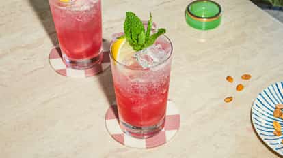 Two Grenadine Fizz cocktails in tall glasses with ice, lemon and mint garnish, served on checkered coasters on a light stone table.