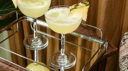 Hawaiian Cosmopolitans in coupe glasses with fresh pineapple slices on a mirrored tray over a wooden surface.