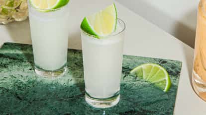 Two Kamekaazi cocktails in frosty shot glasses with fresh lime wedges, served on a green stone board in a bright, modern setting.