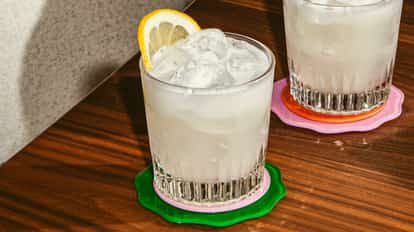 Loneiless Cocktail im Tumbler mit Eis und Zitronenscheibe, helles trübes Getränk auf Holztisch mit bunten Untersetzern.