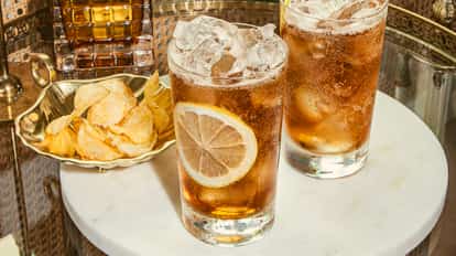 Dos cócteles Long Island Iced Tea con rodajas de limón y hielo sobre mesa de mármol junto a cuenco dorado con patatas fritas.