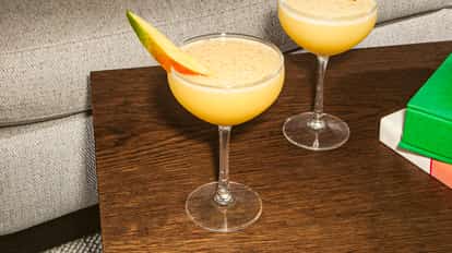 Mango No. 5 cocktail in a coupe glass, creamy yellow with a fresh mango slice garnish, set on a wooden table beside books and a sofa.