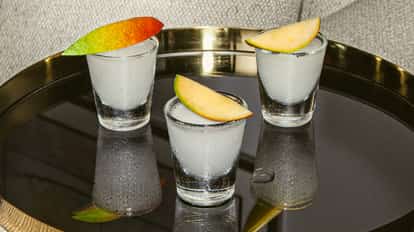 Mango Shooter servido en tres vasos de shot escarchados, cada uno decorado con una rodaja de mango, sobre una bandeja negra brillante.