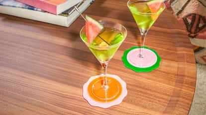 Melon Martini cocktails in chilled martini glasses with green tones, garnished with fresh watermelon slices, set on a wooden table with colorful coasters and books.