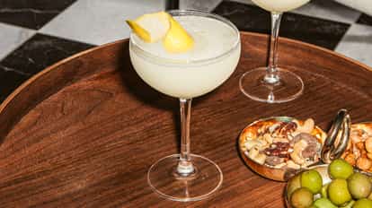 Cócteles Nikita en copas coupe elegantes con superficie clara y espumosa y piel de limón, servidos en una bandeja de madera con frutos secos y aceitunas verdes.