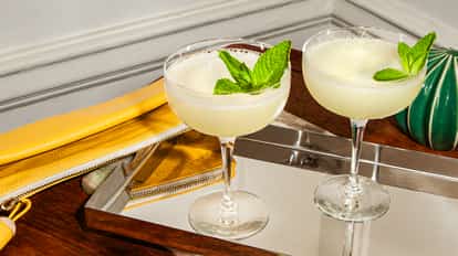 Two coupe glasses of frothy mint-garnished cocktails on a mirrored tray with a yellow clutch and green decor on a wooden surface.