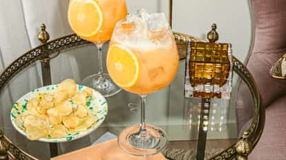 Orang-a-tang cocktail in large stemmed glasses with ice and orange slices, served on a vintage tray beside chips and decorative glassware.