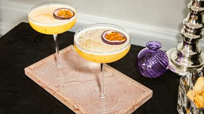 Dos martinis de maracuyá en copas coupe con media maracuyá como adorno, servidos sobre bandeja de mármol rosa.