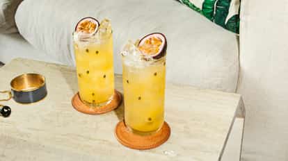 Tall glasses of iced passion fruit cocktails on a marble table, topped with fresh passion fruit halves beside jewelry and a beige sofa.