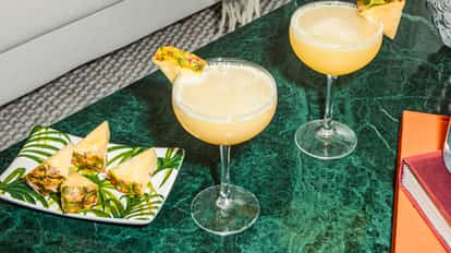 Pineapple Martini cocktails in coupe glasses, topped with frothy pineapple mix and garnished with fresh wedges, served on a green marble table.