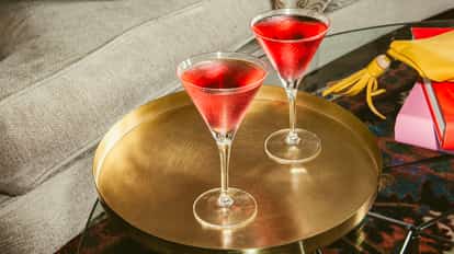 Pink Martini cocktails in chilled martini glasses on a gold tray beside a sofa and colorful books on a glass coffee table.
