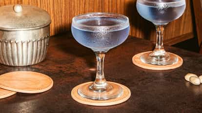 Purple Cosmopolitan served in chilled coupe glasses with frosted sides, placed on leather coasters on a dark bar surface.