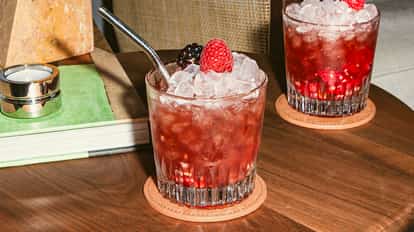 Cóctel Raspberry Blimey servido en un vaso bajo con hielo triturado, de color rojo intenso y decorado con frambuesas frescas y una mora, sobre una mesa de madera.