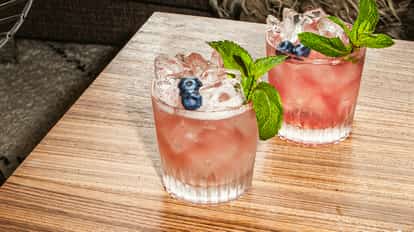 Cócteles Razzle Dazzle en vasos helados, llenos de hielo y decorados con arándanos frescos y menta, colocados sobre una mesa de madera.