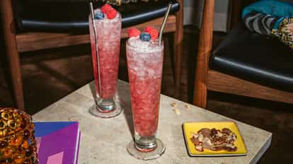 Coquetel Russian Spring Punch em um copo alto com gelo triturado, cor rosa e decorado com framboesas e um mirtilo, sobre uma mesa de pedra.