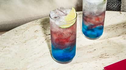 Sparkler cocktail in tall glass with layered blue and red hues, ice and lime wedge, served on a light stone table.