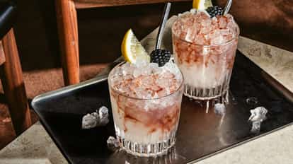 Vodka Bramble Cocktail im Tumblerglas mit Crushed Ice, rosafarbenen Wirbeln und Garnitur aus Zitrone und Brombeere, auf einem Metalltablett serviert.