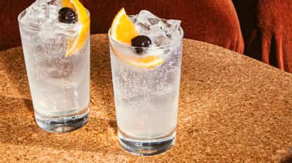 Cóctel Vodka Collins en vaso alto con burbujas claras, cubos de hielo y una decoración de rodaja de naranja y cereza oscura sobre una mesa de corcho.