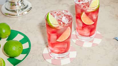 Dos cócteles vodka cranberry con hielo y lima sobre posavasos estampados junto a un plato de limas frescas en superficie clara.