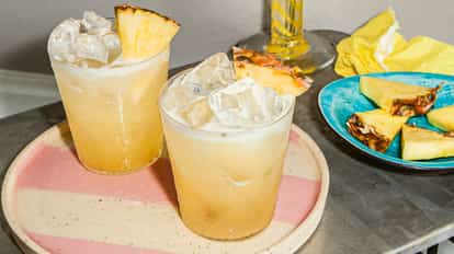 Vodka Fizz cocktails in ice-filled glasses with pineapple garnishes on a pink-striped plate beside a dish of fresh pineapple slices.