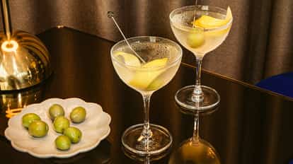 Two vodka martini cocktails with lemon peel and green olives on a dark glossy table beside a plate of fresh green olives.