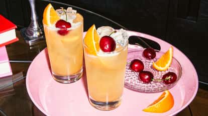 Yellowhammer cocktails in tall ice-filled glasses garnished with orange slices and cherries on a pink tray beside a small plate of cherries and orange slices.