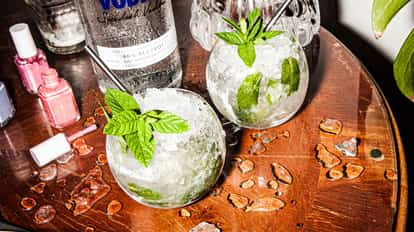 Two refreshing mint juleps served in round glasses filled with crushed ice and fresh mint leaves, placed on a wooden table with a vodka bottle, scattered ice, and nail polish bottles nearby.