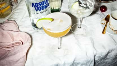 Apple Strudel Martini in a coupe glass garnished with an apple slice beside an Absolut Vodka bottle on white background.