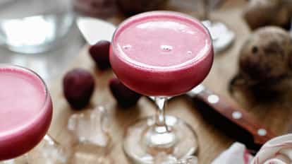 Beets Martini in a coupe glass with a frothy head on a wooden board beside beet halves and ice cubes