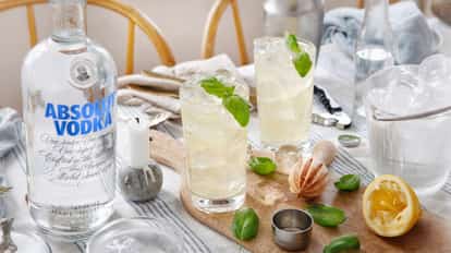Two highball glasses of basil cocktail with basil leaves and ice on a board beside an Absolut vodka bottle.